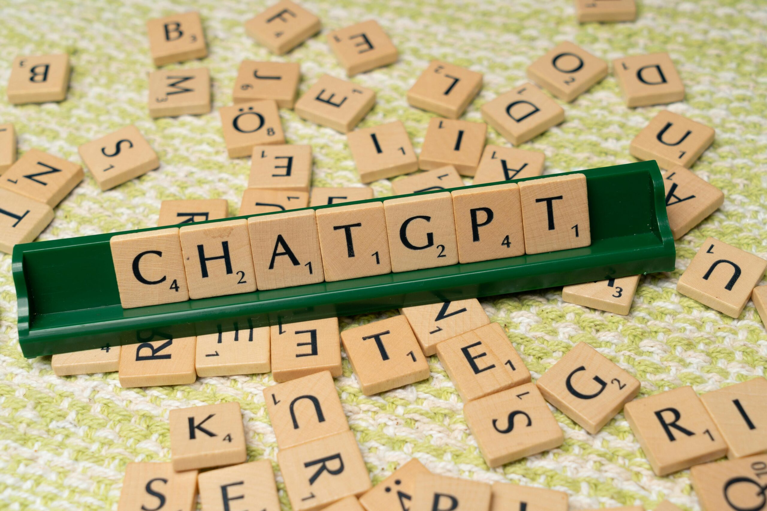 Lettres en bois du jeu Scrabble formant le mot 'CHATGPT' sur un support vert, avec des lettres éparpillées en arrière-plan.