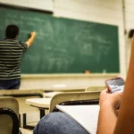 Un étudiant au premier plan utilise discrètement son smartphone pendant qu'un professeur écrit au tableau noir dans une salle de classe. Des bureaux et des chaises sont visibles, et l'étudiant tient un cahier ouvert.