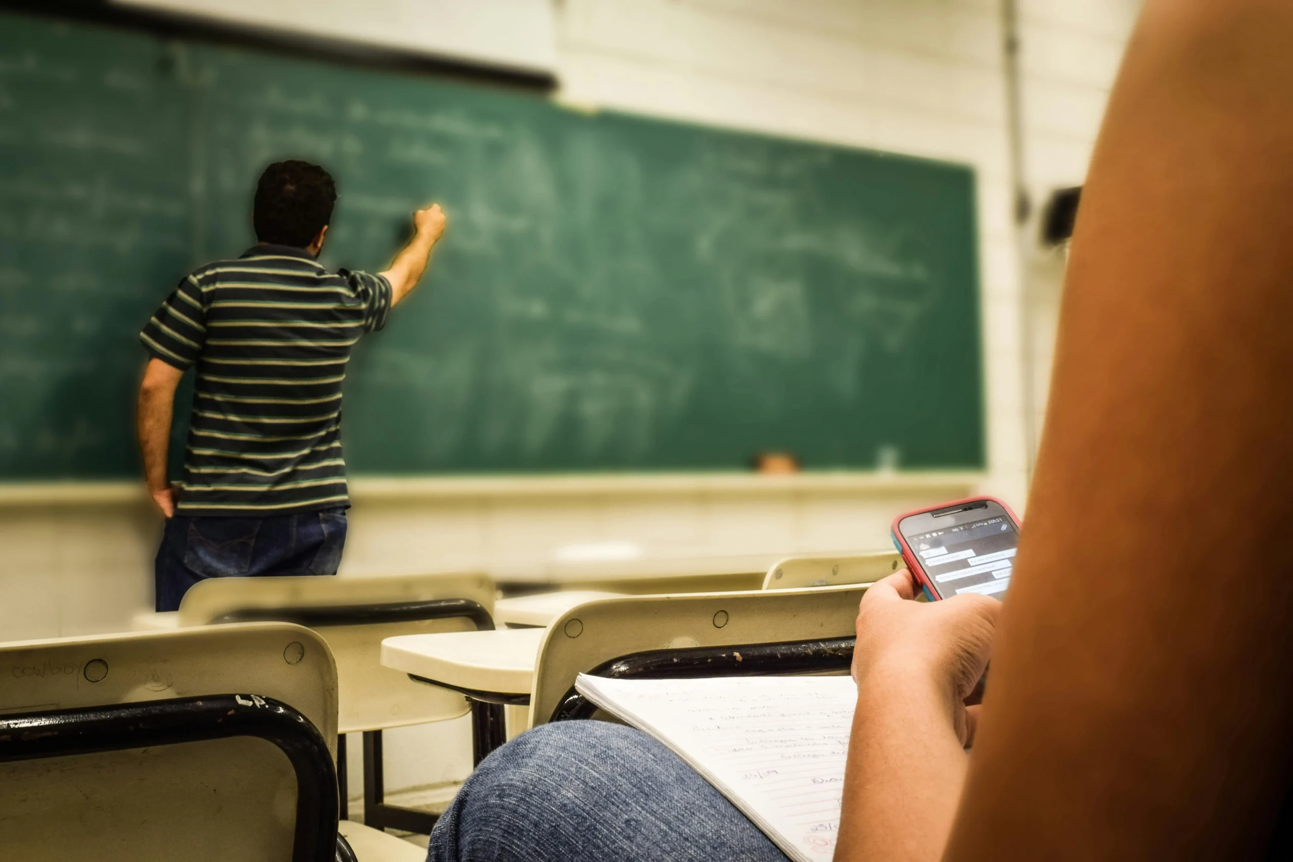 Un étudiant au premier plan utilise discrètement son smartphone pendant qu'un professeur écrit au tableau noir dans une salle de classe. Des bureaux et des chaises sont visibles, et l'étudiant tient un cahier ouvert.