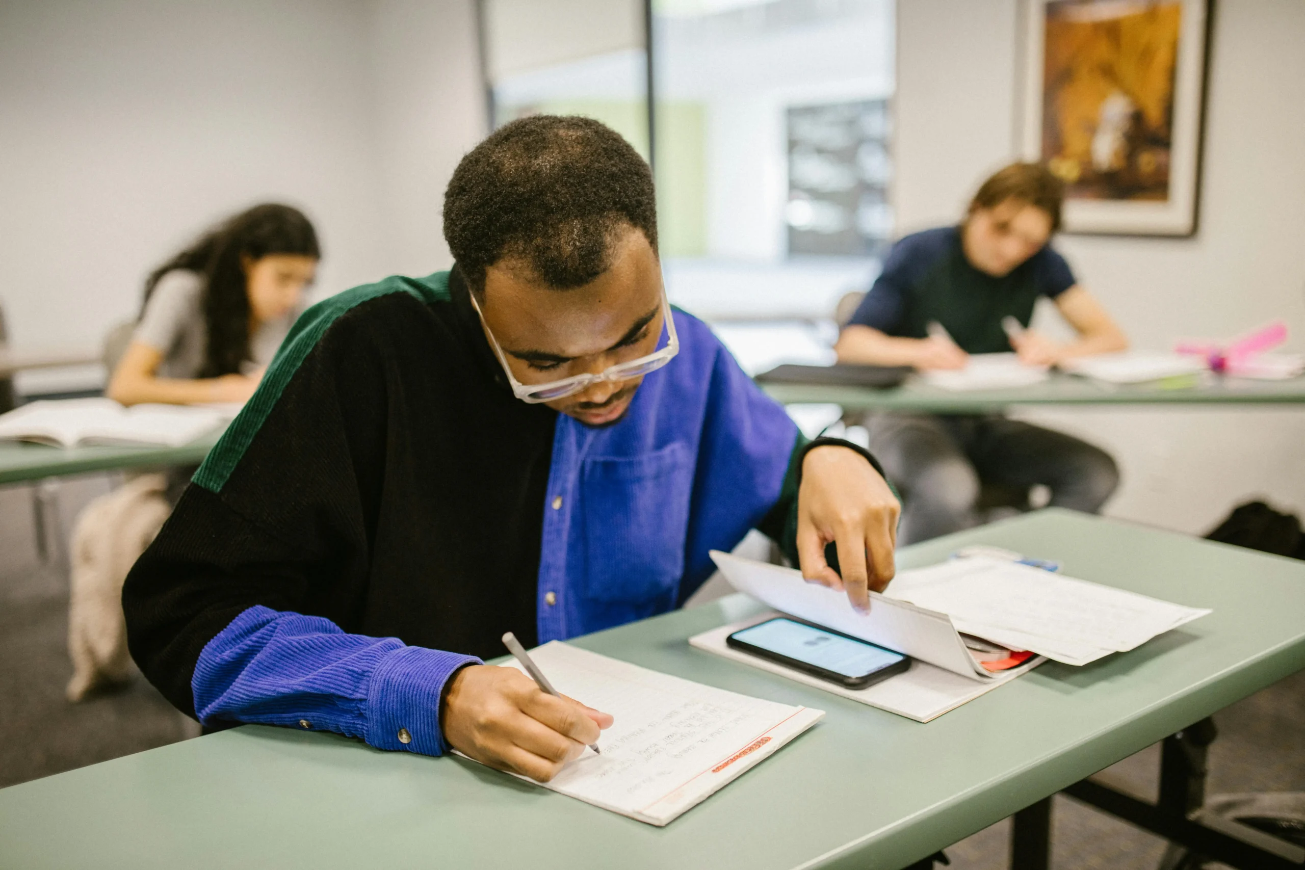 Un étudiant portant une chemise multicolore est entrain de tricher. Un smartphone est posé à côté de lui, tandis que d'autres étudiants travaillent en arrière-plan.