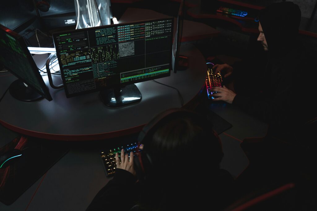 Deux individus travaillant sur des ordinateurs dans un environnement sombre, tapant du code sur des claviers rétroéclairés, avec des écrans affichant des lignes de données techniques.