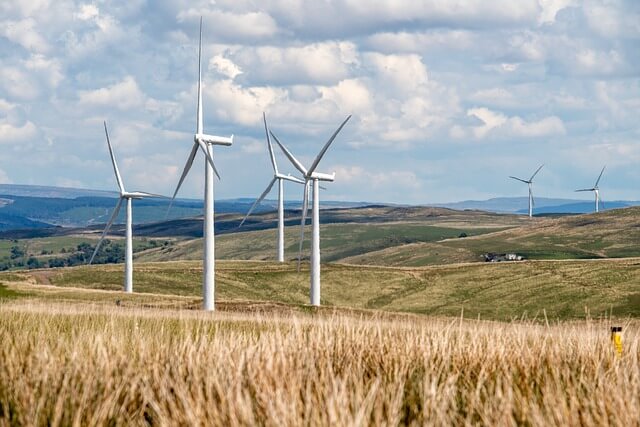 Éoliennes dans une région rurale avec des champs dorés et des collines sous un ciel nuageux.
