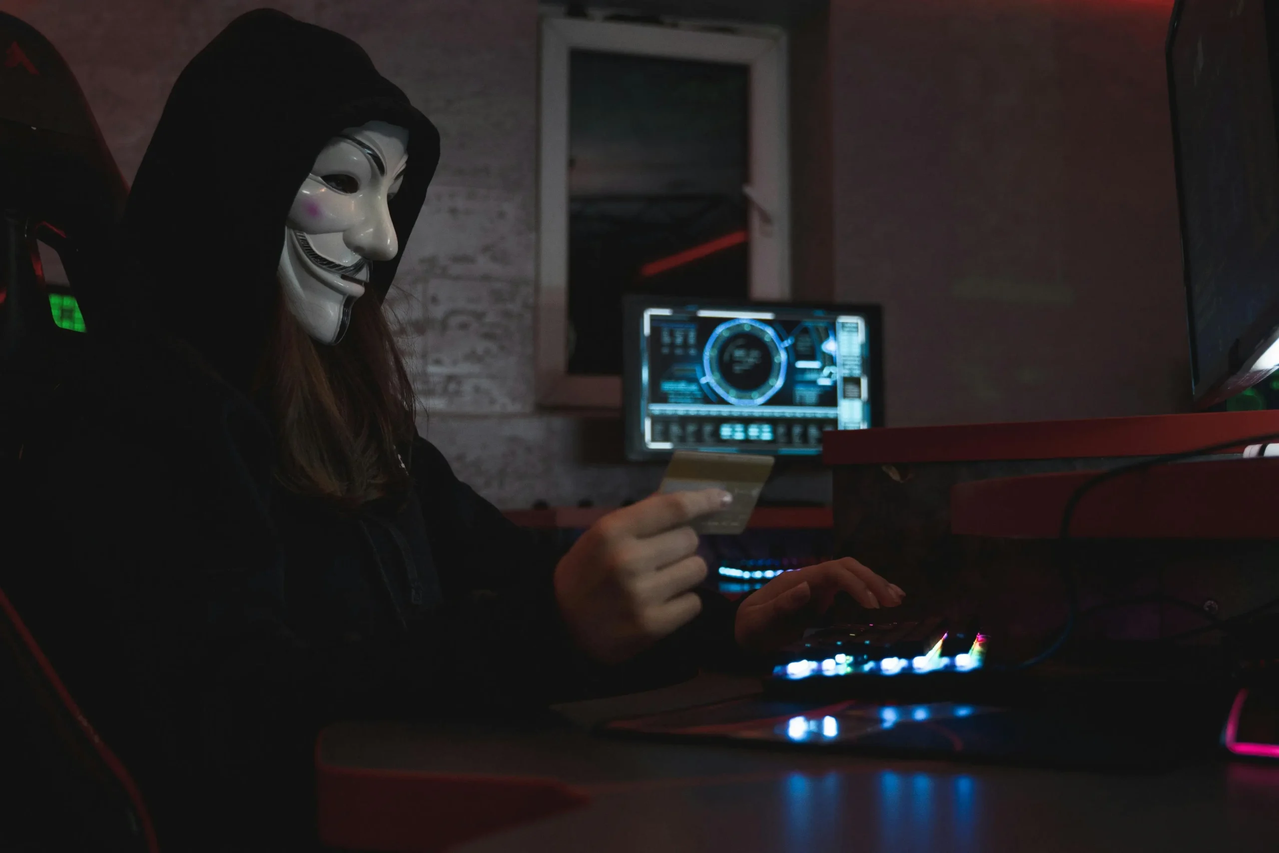 Personne masquée avec un masque de Guy Fawkes et un hoodie tenant une carte bancaire devant un ordinateur dans un environnement sombre.